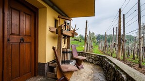 A stone-floored porch with two wooden chairs, a wooden door, and a rustic outdoor setting with a fence and greenery.