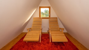 A small attic room with two wooden chairs, a red carpet, and a window with a view of greenery.