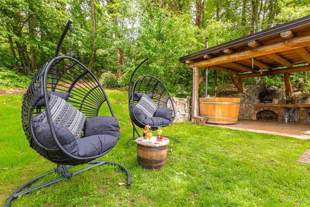 Dos sillas colgantes negras con cojines, un barril de madera, y una chimenea de piedra bajo una pérgola de madera en un jardín exuberante.