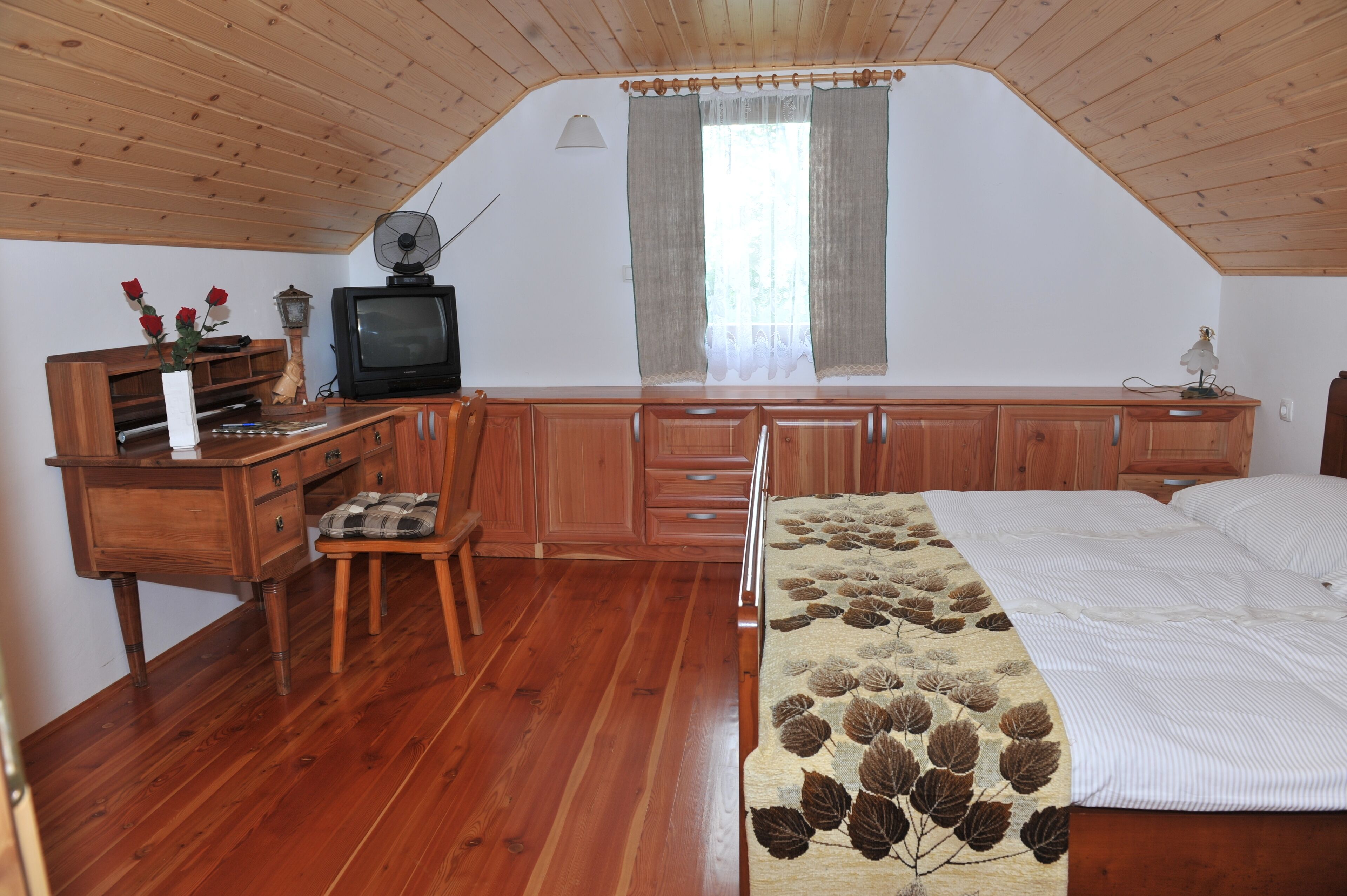 Une chambre avec un plafond en bois, un lit recouvert d’une couvre-lit à motifs de feuilles, un bureau avec une télévision et une chaise.