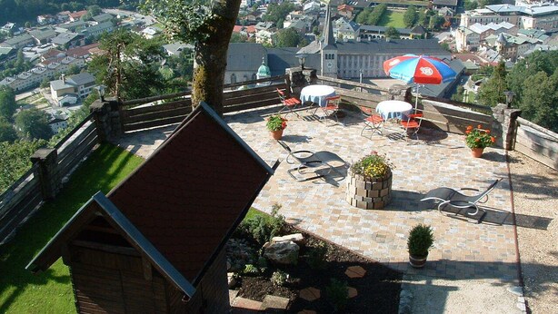 Una terraza en la azotea con mobiliario exterior, vistas a un pueblo y un edificio pequeño con techo rojo.