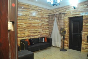 A room with a marble-patterned wall, a black sofa with cushions, a standing fan, and a door. - Tobby Royal Suites (Lagos)