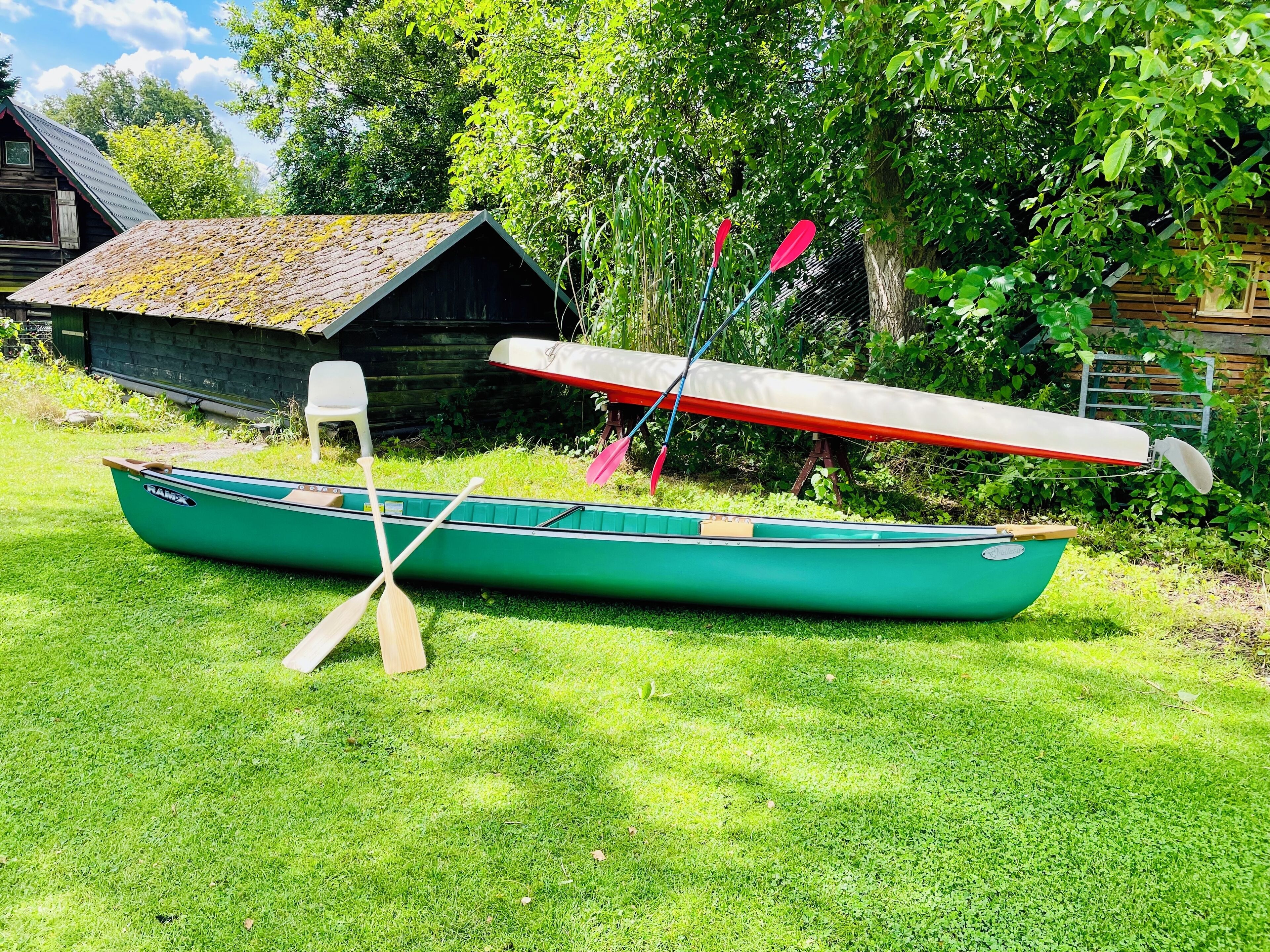 Una canoa verde con remos y un remo rojo apoyado contra un cobertizo de madera en una zona cubierta de césped.