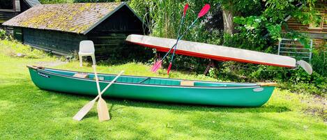Una canoa verde con remos y un remo rojo apoyado contra un cobertizo de madera en una zona cubierta de césped.