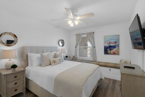 Une chambre à coucher comprenant un lit, des tables de chevet, un ventilateur de plafond, une fenêtre avec des rideaux, un miroir et des œuvres d’art accrochées au mur.