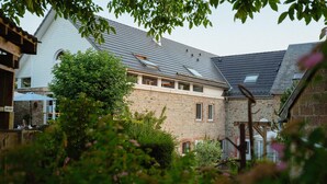 A stone building with a sloped roof, a wooden pergola, and a garden area with bushes and flowers. - Group Getaway in Amel (Amel)