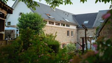 A stone building with a sloped roof, a wooden pergola, and a garden area with bushes and flowers.