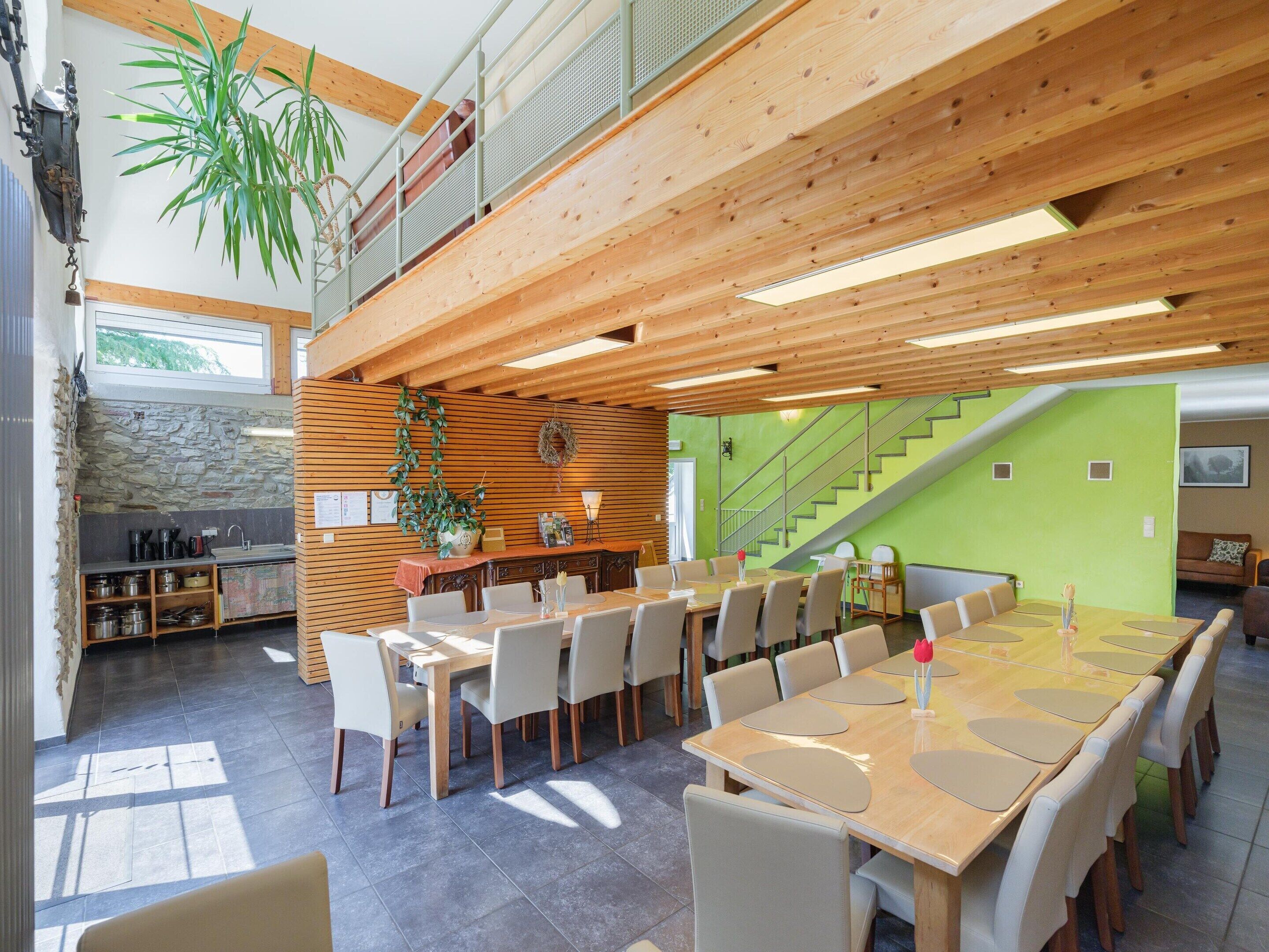 A spacious dining area with a long wooden table and chairs, a kitchen area with stone wall and wooden cabinets, and a staircase leading to an upper level.
