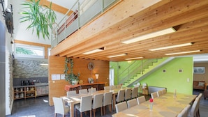A spacious dining area with a long wooden table and chairs, a kitchen area with stone wall and wooden cabinets, and a staircase leading to an upper level. - Group Getaway in Amel (Amel)