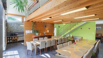 A spacious dining area with a long wooden table and chairs, a kitchen area with stone wall and wooden cabinets, and a staircase leading to an upper level.