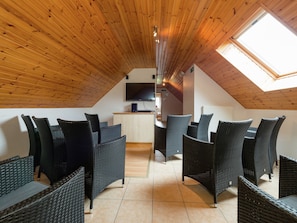 A high-ceilinged room with wooden beams, a dining area with wicker furniture, and a TV mounted on the wall.