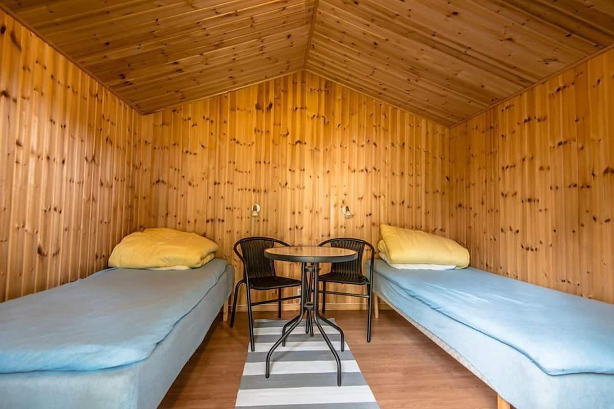 A small, cozy room with wooden walls and a sloped ceiling. There are two single beds with blue bedding and yellow pillows. A small round table with two chairs is placed in the center.