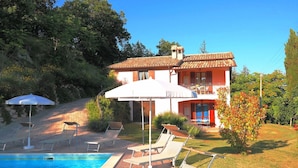 Ein zweistöckiges Haus mit Swimmingpool, Liegestühlen und einem weißen Sonnenschirm.