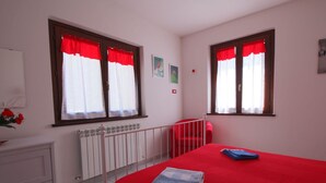 Ein Schlafzimmer mit roter Bettdecke, zwei Fenstern mit roten Vorhängen, einem weißen Heizkörper und einer weißen Kommode.