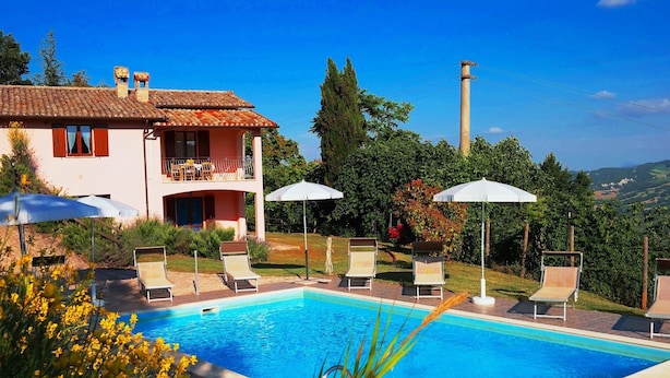 Ein Poolbereich mit Liegestühlen und Sonnenschirmen, ein zweistöckiges Gebäude mit Balkon sowie Blick auf die umliegende Landschaft.