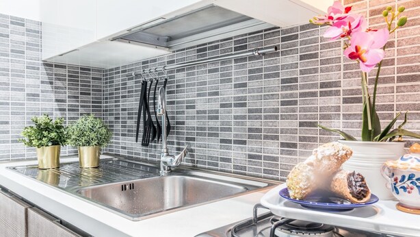 A modern kitchen with a stainless steel sink, a gas stove, and a tiled backsplash. There are two potted plants on the counter and a vase with flowers on the stove.