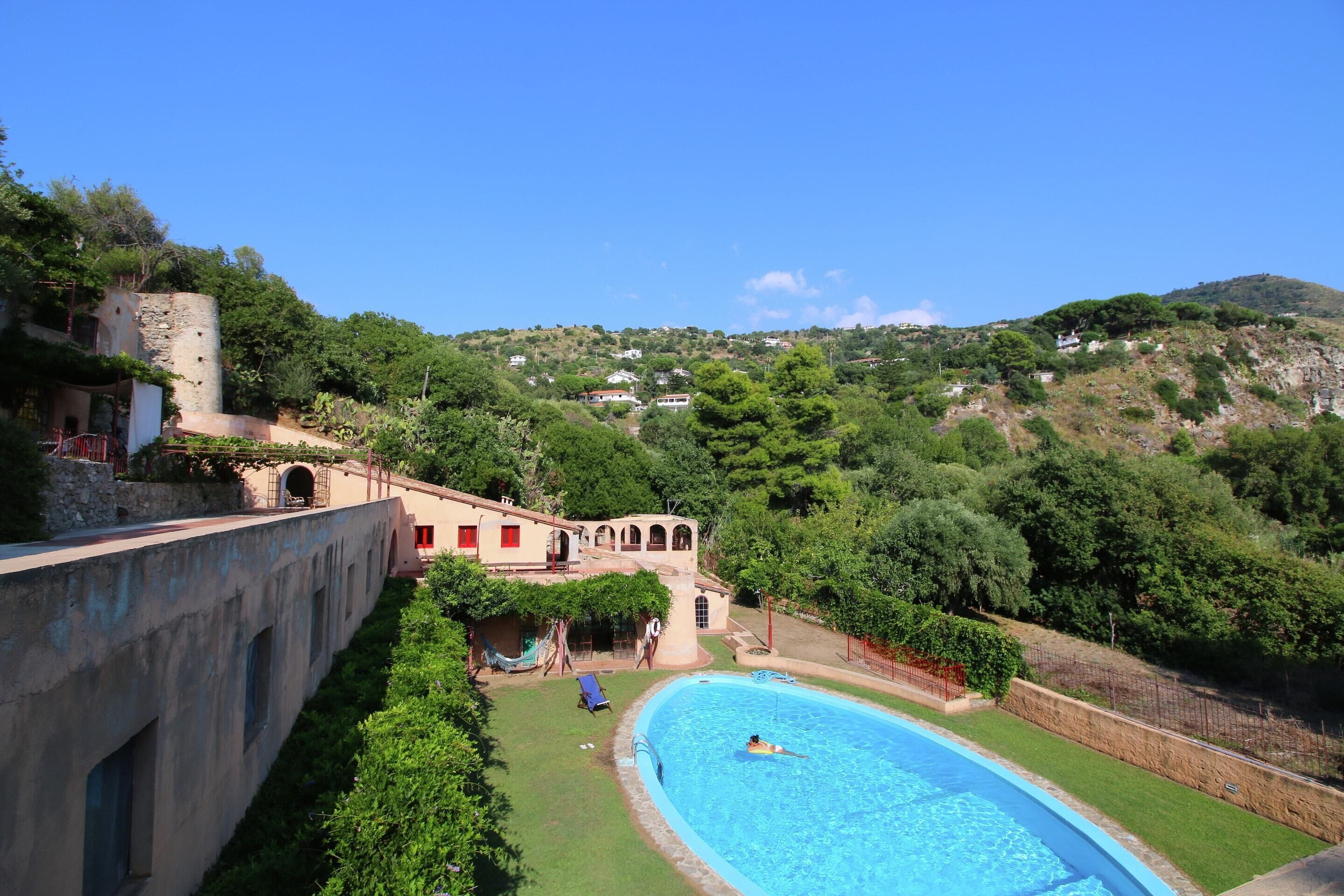 Une piscine avec une personne dans l’eau, entourée de verdure et avec une colline en arrière-plan.