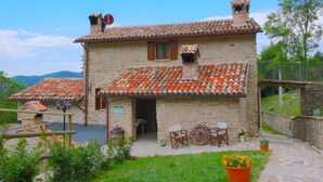 A stone building with a tiled roof, a wooden door, and a small porch with two chairs and a table. - Farmhouse in Marche With Pool and Views (Mercatello sul Metauro)