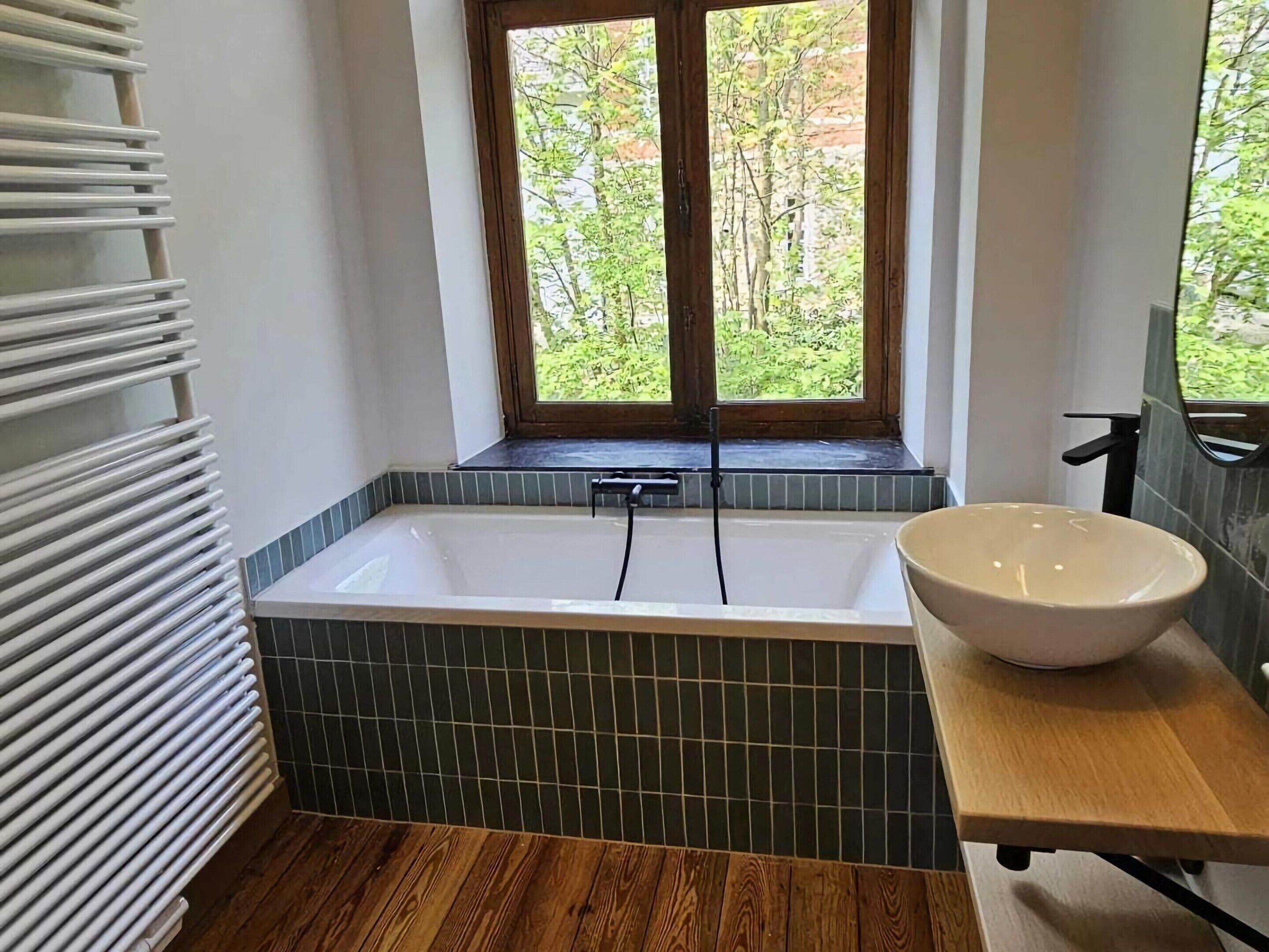 A modern bathroom with a freestanding bathtub, a wooden vanity, and a window with a view of greenery.