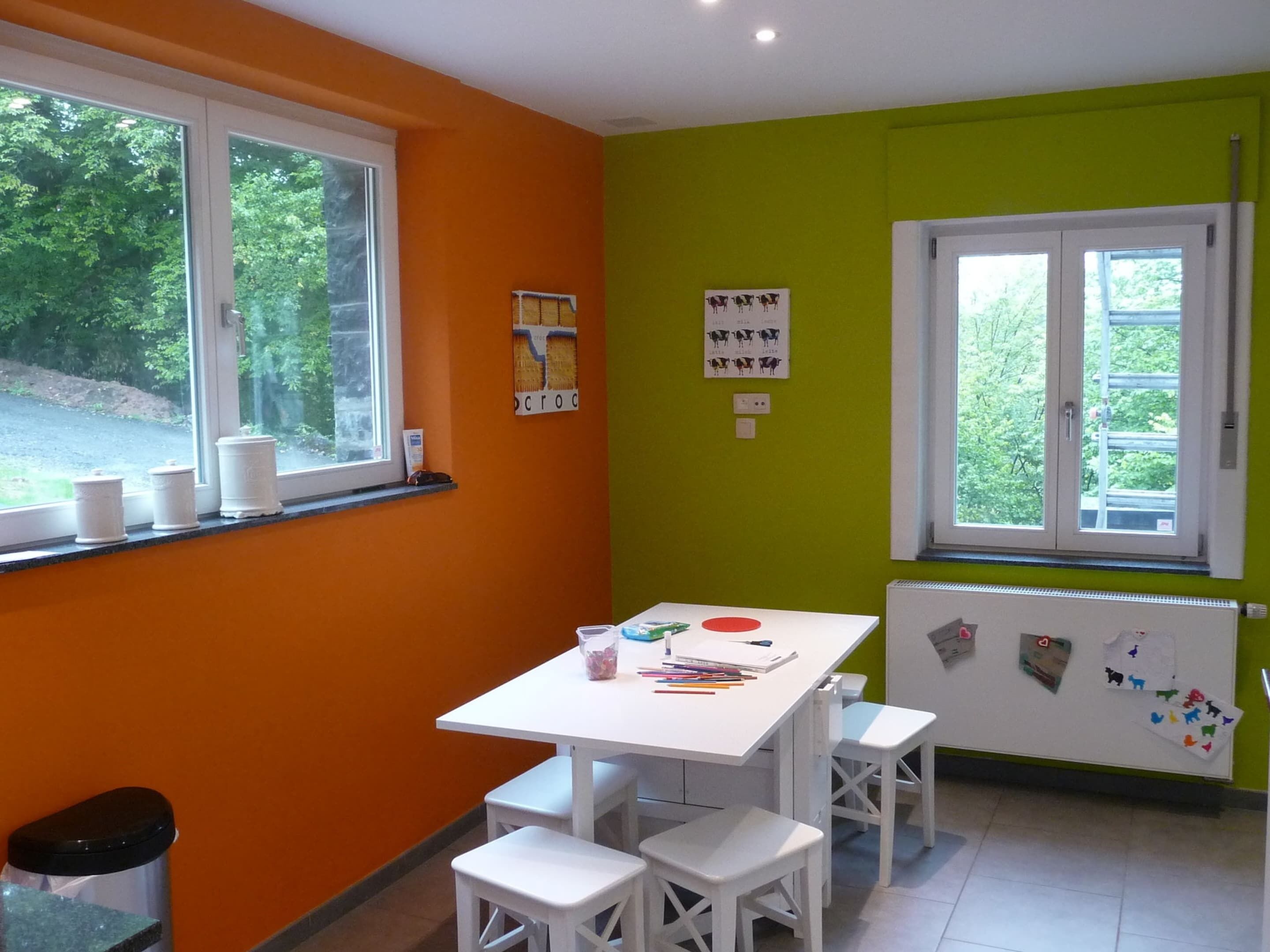 A classroom with a white table and benches, a window with a view of greenery, and educational posters on the walls.