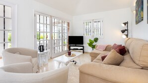Ein großzügiges Wohnzimmer mit einem beigen Sofa, einem weißen Sessel und einem Couchtisch. Ein Flachbildfernseher ist an der Wand befestigt und es gibt große Fenster mit weißen Rahmen.
