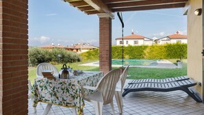 Une terrasse couverte avec une table dressée pour deux personnes, un fauteuil suspendu et une vue sur une piscine et des maisons au loin.