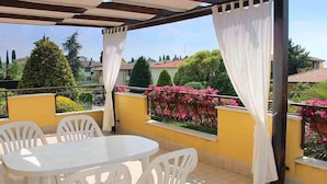 A covered outdoor seating area with white plastic chairs and a table, surrounded by yellow walls and a flower box with pink flowers. - Holiday Home in Lazise Near Gardaland (Lazise)