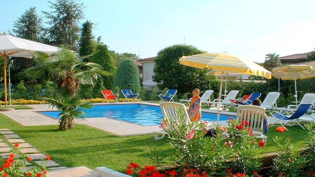 A pool area with lounge chairs and umbrellas, surrounded by greenery and a brick wall. - Holiday Home in Lazise Near Gardaland (Lazise)