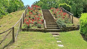 Uma escadaria de pedra com corrimão de madeira, rodeada por vegetação exuberante e flores vermelhas.