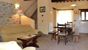 Uma sala de estar acolhedora com parede de pedra, vigas de madeira, um sofá, uma mesa e um espaço de jantar com cadeiras e um banco.