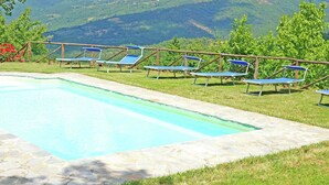 Piscina retangular com borda de pedra, rodeada por um gramado com espreguiçadeiras azuis e cerca de madeira.