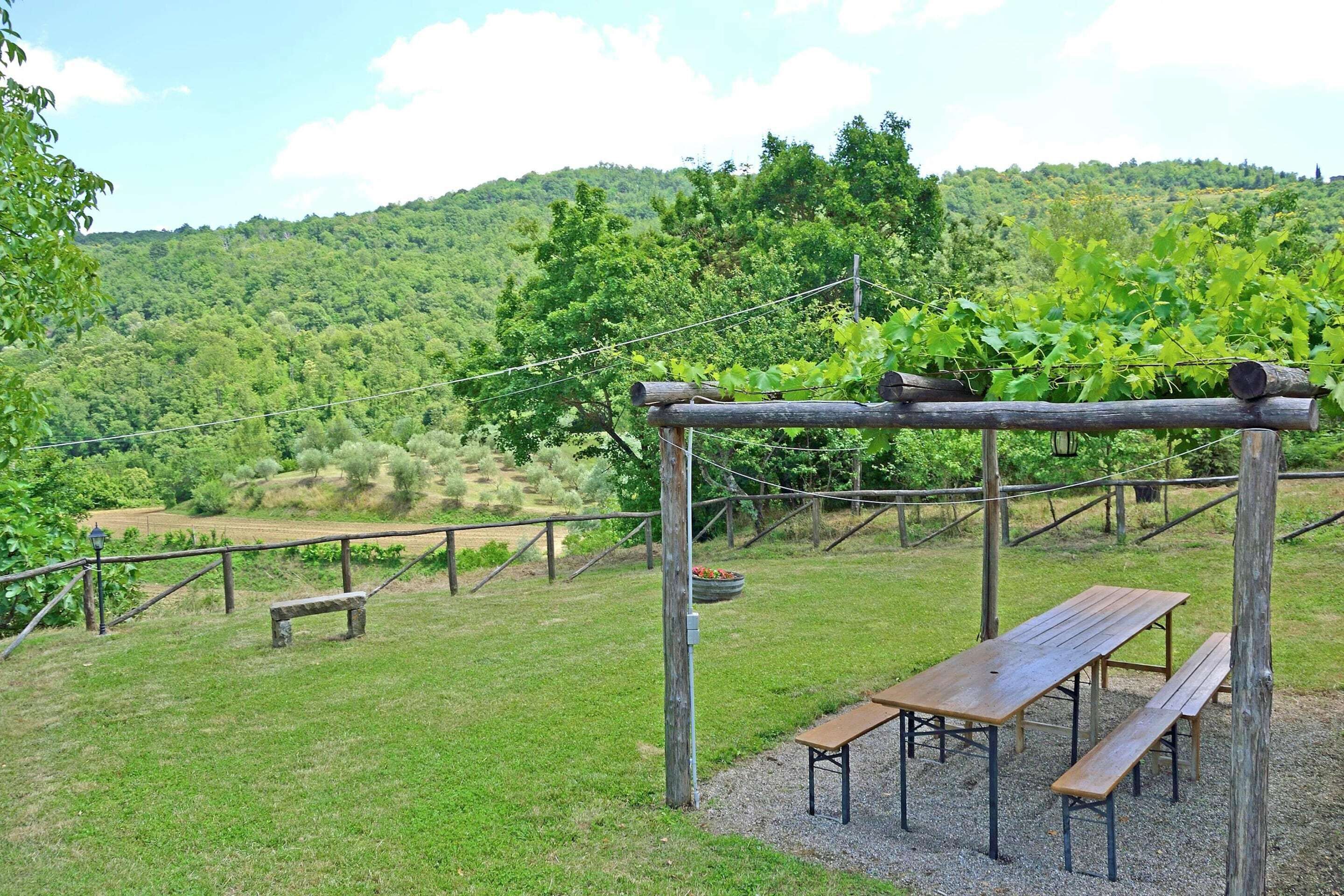 A rustic outdoor seating area with wooden benches and a pergola, surrounded by lush greenery and hills.
