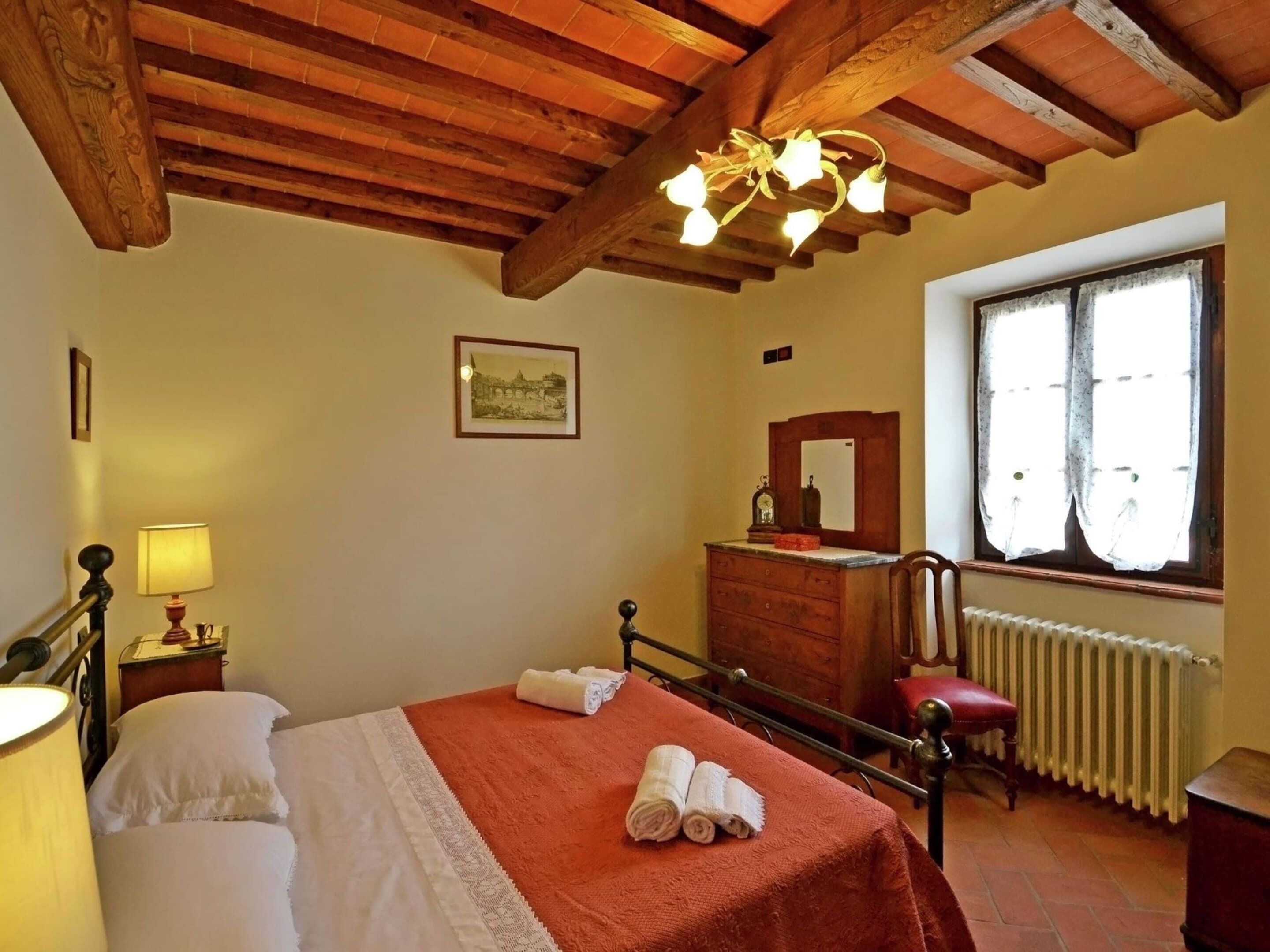 A bedroom with a wooden ceiling, a bed with a red blanket, a nightstand with a lamp, a dresser, a mirror, and a window with curtains.