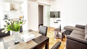 A modern living room with a sofa, a dining table with a vase of flowers, and a TV mounted on the wall. - Luxury Apartment With Private Balcony in Quiet Area (Dubrovnik)