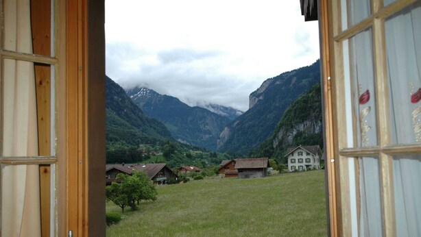 A view from inside a room with wooden walls and a window, looking out at a mountain landscape with houses and a grassy field. - Pristine Home in a Charming Village, Large Grassy Sunbathing Area, View of the Mönch and Jungfrau (Wilderswil)