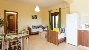 A modern kitchen with a refrigerator, a dining area with a table and chairs, a sofa with cushions, and a window with curtains. - New Small Apartmentcomplex in Village of Prinès near Rethymnon (Rethymno)