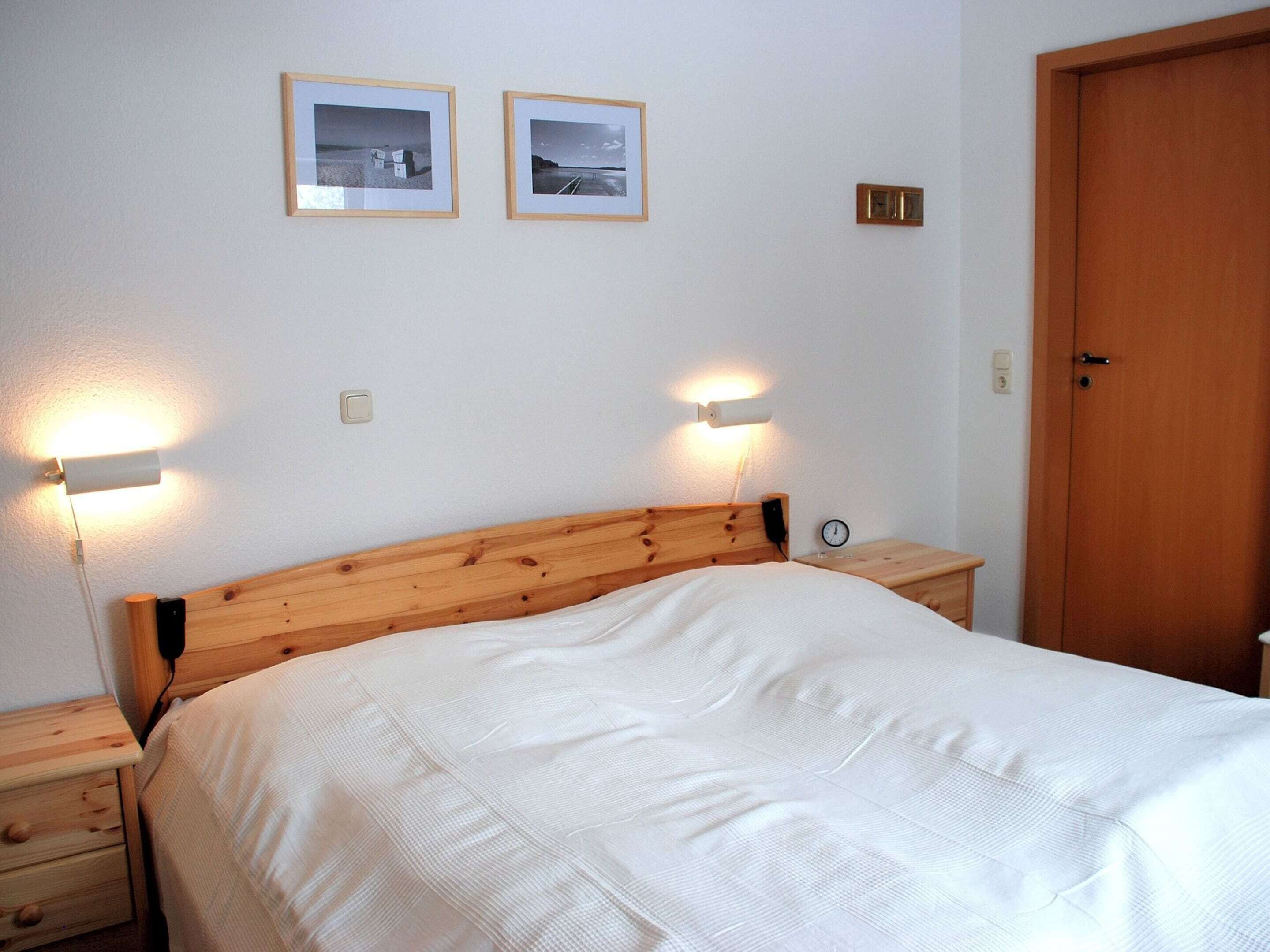 Een slaapkamer met een houten hoofdeinde, een bed met wit beddengoed, twee nachtkastjes met lampen, en twee ingelijste foto’s aan de muur.