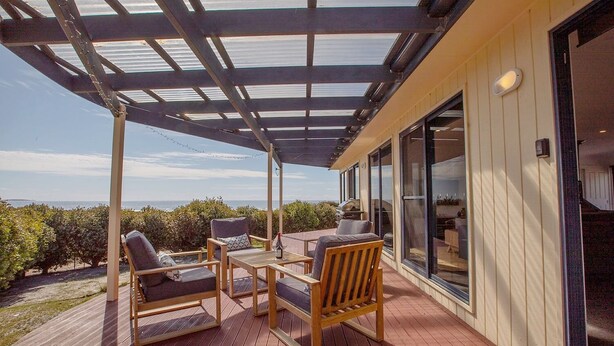 Une terrasse extérieure couverte, aménagée avec du mobilier en bois, offrant une vue sur la mer et donnant accès à l’intérieur par une porte vitrée.