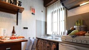 A kitchen with a stove, sink, and various cooking utensils. There is a window with blinds, a wooden shelf with pots, and a bottle on the counter. - Tranquil Holiday Home in Houffalize With Barbecue (Houffalize)