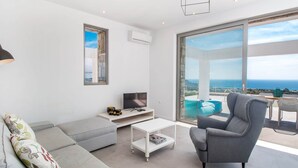 A modern living room with a grey sofa, a grey armchair, a white coffee table, and a flat-screen TV. There is a large window offering a view of the sea. - Sea View Villa in Kalythies with Private Pool near 3 Beaches (Rhodes)