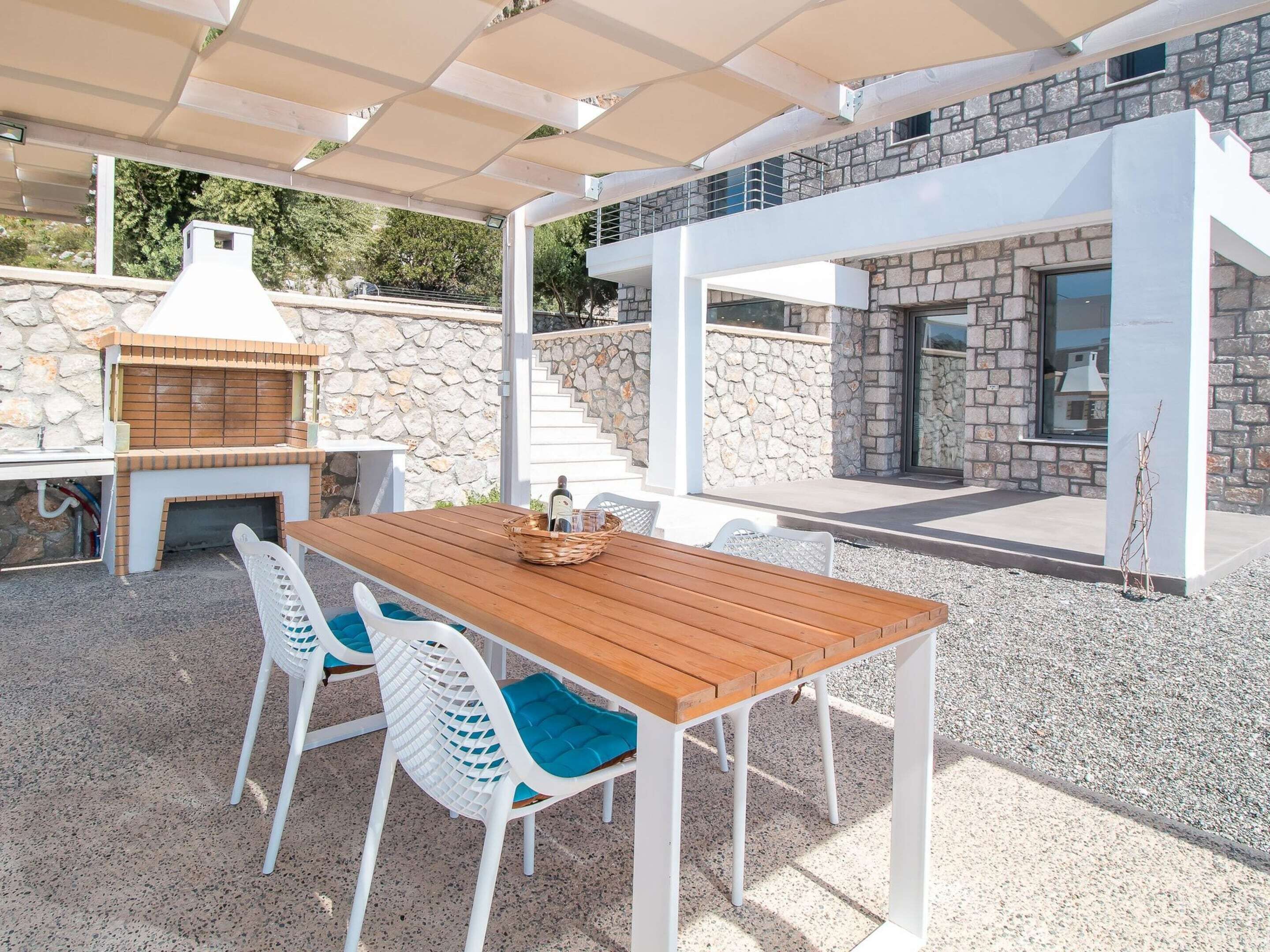 Un patio al aire libre con mesa de madera y sillas blancas, una parrilla y un muro de piedra.