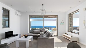 A modern living room with a grey sofa, white coffee table, and a flat-screen TV. There is a large window with a view of the sea. - Sea View Villa in Kalythies with Private Pool near 3 Beaches (Rhodes)