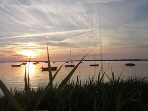A sunset over a calm body of water with several boats anchored, surrounded by grassy areas.