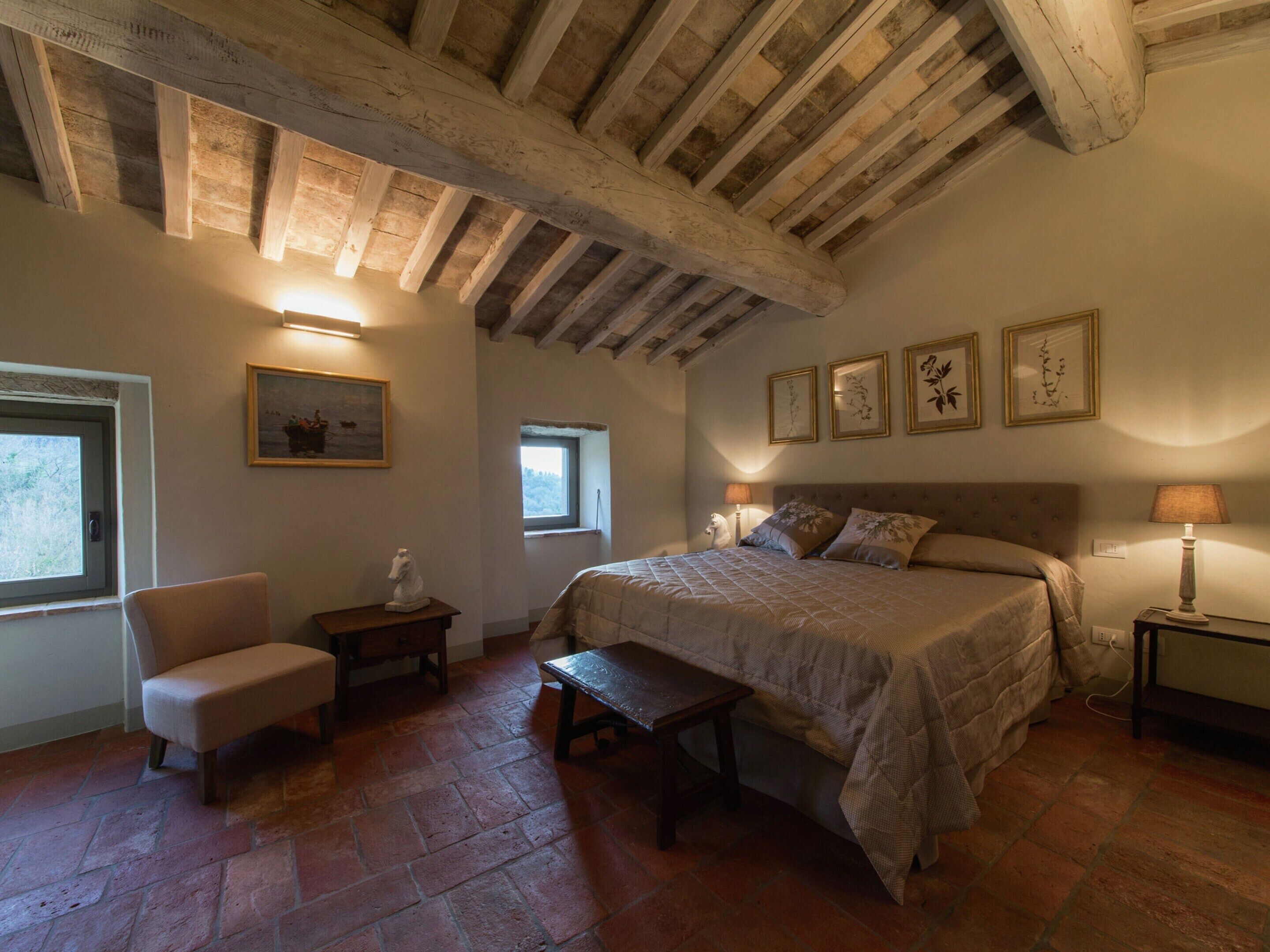 A bedroom with a large bed, a chair, a small table, and a bench. There are framed pictures on the wall and a window with a view of trees.