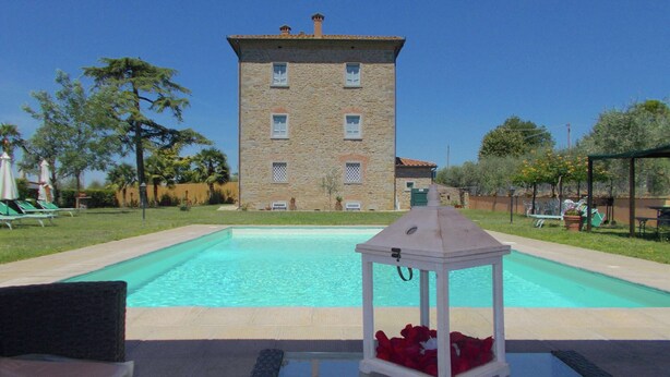 Une piscine avec un bâtiment en pierre en arrière-plan, une table en verre avec une lanterne et du mobilier d’extérieur.