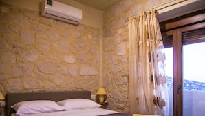A bedroom with a stone wall, a bed with white linens, a nightstand with a lamp, and a view of a mountainous landscape through a window. - Luxurious Villa in Bali Crete With Private Pool (Mylopotamos)