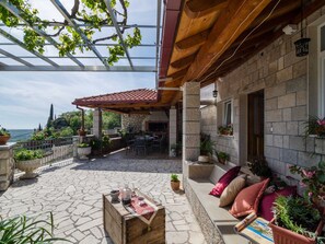 Eine überdachte Terrasse mit Steinboden, einem Holztisch mit Flasche und Gläsern sowie einer Sitzecke mit Kissen und Topfpflanzen.