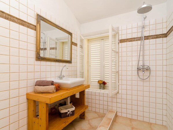 Une salle de bain avec un meuble-lavabo en bois, un évier, un miroir, une fenêtre avec des volets et une douche.