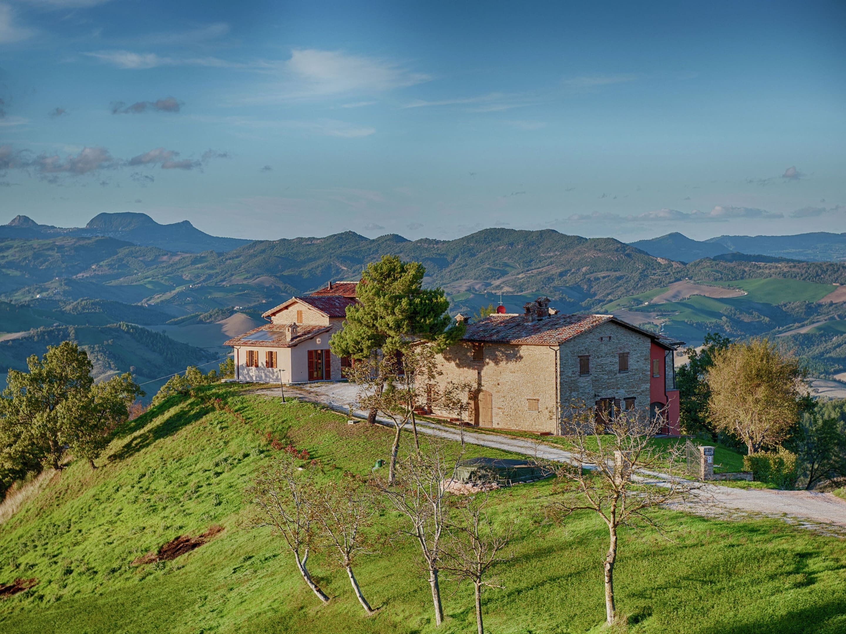 A stone house with a red roof on a hillside, surrounded by green fields and mountains.