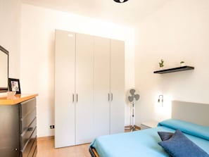 A bedroom with a bed, a wardrobe, a desk, a fan, and a shelf with plants.